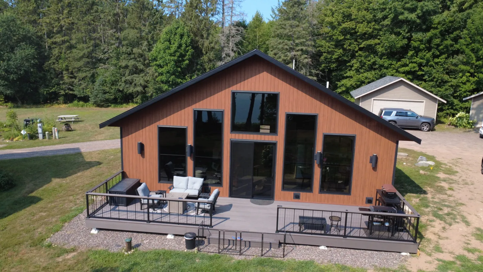 Aerial view of a modern lakefront cabin with a wood-toned exterior, black trim and a wide deck facing the water.